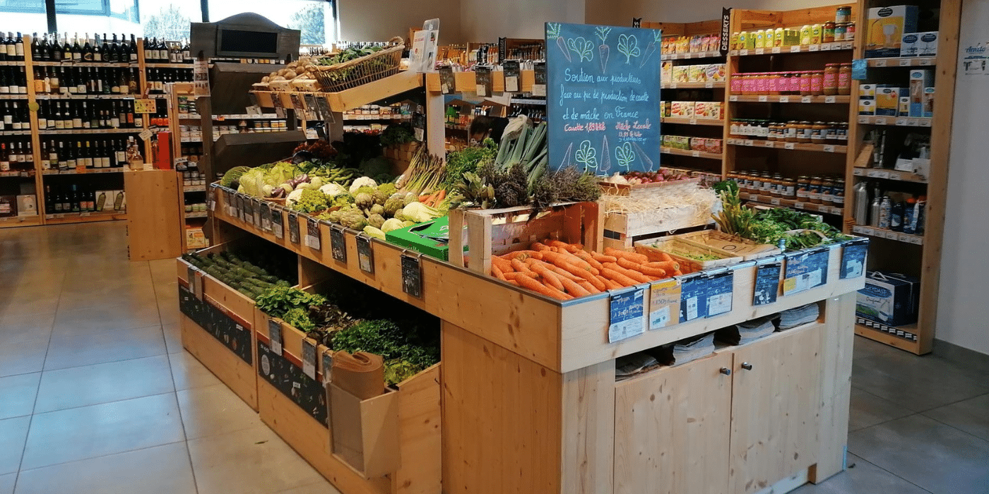 Étal de produits frais en bois dans un magasin Biocoop : Une section rustique avec des étagères en bois, remplie de légumes frais, met en avant l'authenticité et le soutien aux producteurs locaux. Une ardoise signale l'importance du commerce équitable et de la production française, illustrant l'engagement de Biocoop pour des pratiques de consommation respectueuses de l'environnement et orientées zéro déchet. Ce cadre chaleureux et naturel souligne la mission de Biocoop de promouvoir des modes de vie plus durables.