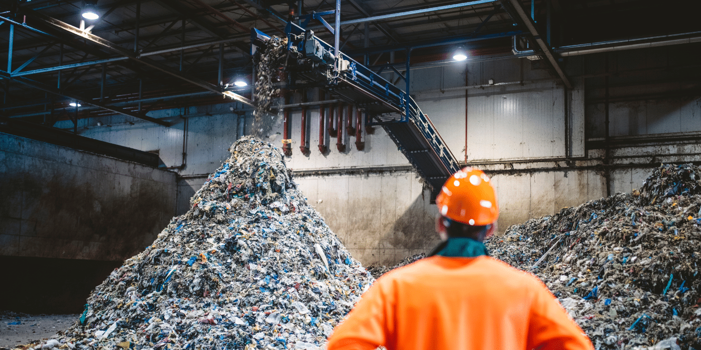 Un ouvrier portant un casque orange observe un tas massif de déchets dans une usine de traitement, où des machines déversent des matériaux recyclables ou non triés sur une pile.