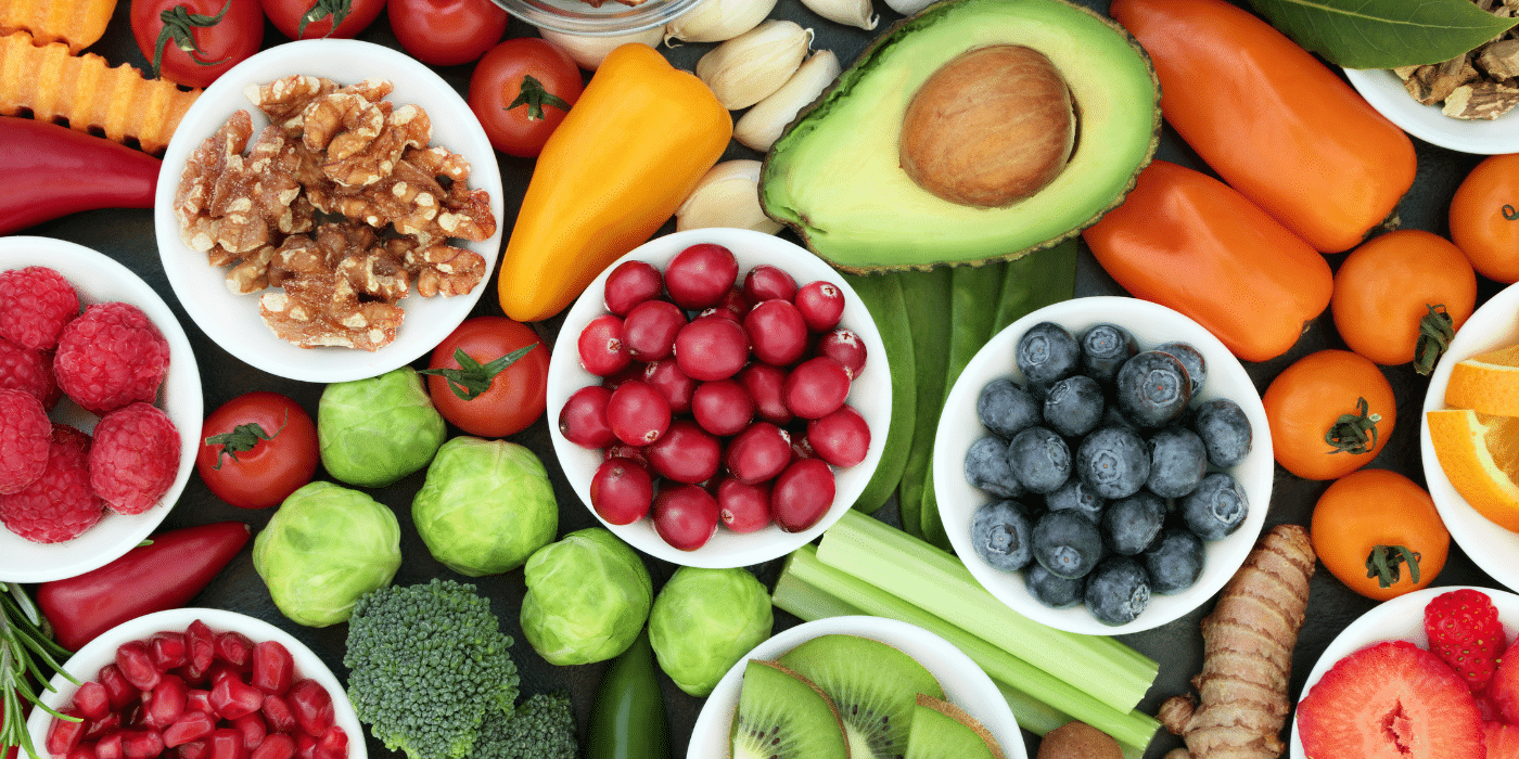 L'image présente un assortiment coloré de fruits, légumes, et noix, soigneusement disposés en vue rapprochée. On peut y voir des bols contenant des baies rouges comme les framboises, les canneberges et les myrtilles, ainsi que des ingrédients riches en nutriments comme les noix et les légumes verts. Autour de ces bols, d'autres aliments sont disposés : avocat, tomates cerises, choux de Bruxelles, céleri, brocoli, poivrons jaunes et oranges, morceaux d'orange, gingembre, et kiwi. Chaque élément semble frais et vibrant, suggérant une alimentation saine et équilibrée. Les couleurs variées, du rouge vif au vert intense en passant par le jaune et l'orange, créent une composition visuelle attrayante et harmonieuse. Cette image évoque l'idée de bien-être, de nutrition équilibrée, et de mode de vie sain, mettant en avant des aliments riches en vitamines, fibres, et antioxydants.