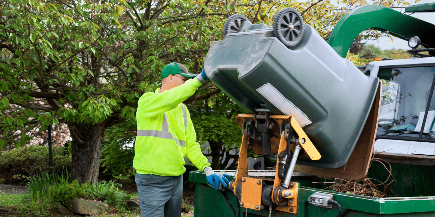 Un employé en tenue de sécurité, portant une veste fluorescente et une casquette verte, vide une poubelle verte dans un camion de collecte des déchets dans un environnement de jardin verdoyant.