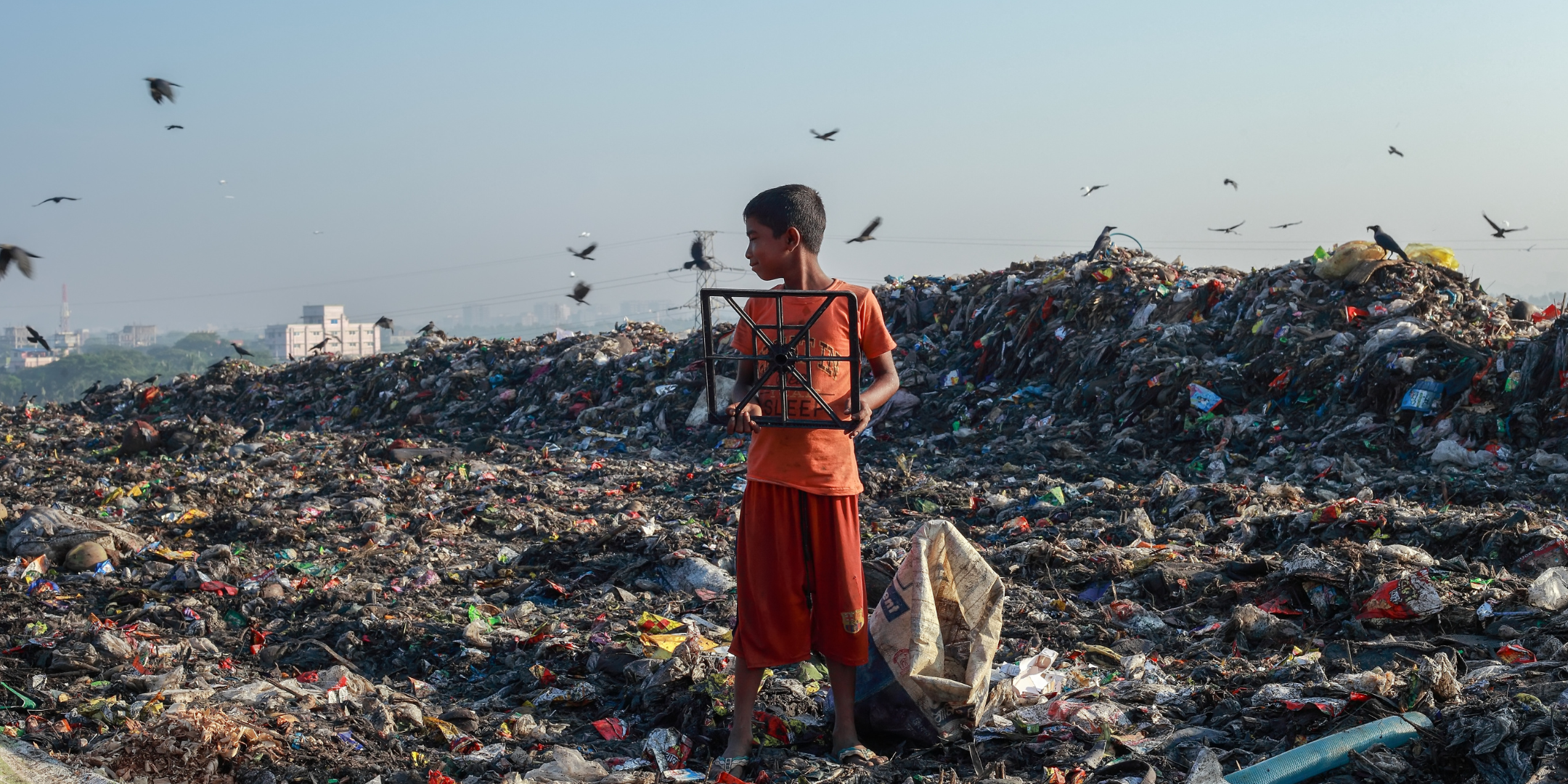 Un enfant portant un cadre métallique dans une décharge de déchets, entouré de montagnes de détritus. Cela illustre l'impact environnemental des déchets électroniques dans certaines régions et l'importance de trouver des solutions durables pour gérer ces déchets, notamment à travers le recyclage.