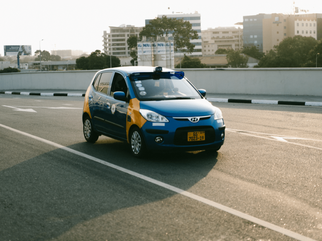 Petit taxi bleu et jaune circulant sur une route urbaine avec des immeubles en arrière-plan.