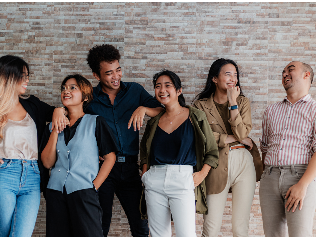 Groupe de six jeunes adultes souriants debout devant un mur en briques, ambiance détendue et conviviale