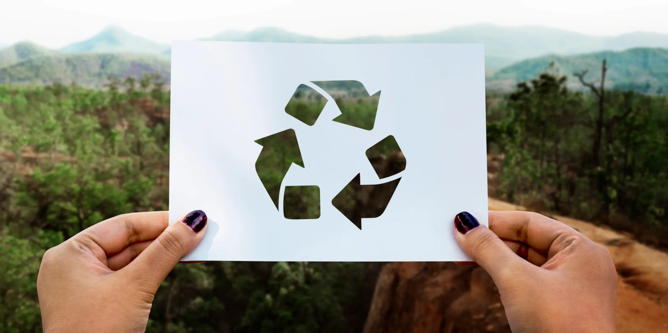 Symbole de recyclage découpé dans une feuille tenu face à un paysage naturel, illustrant les gestes écologiques pour réduire son impact environnemental