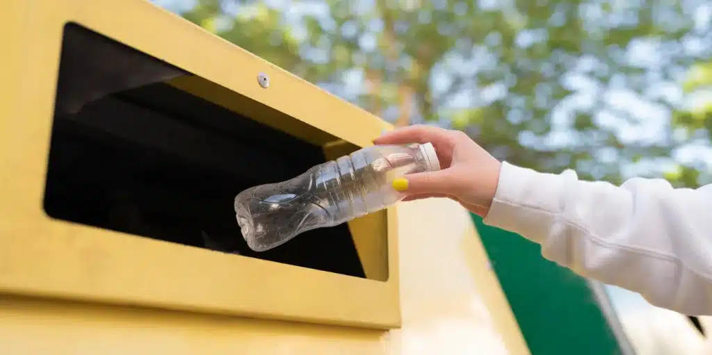 Main déposant une bouteille en plastique vide, bouchon vissé, dans un conteneur jaune